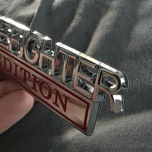 以下が含まれることがあります： 「FIGHTER」の文字が浮き彫りになったクロム仕上げのエンブレム。赤い文字で「EDITION」と書かれています。グレーの背景にエンブレムが配置されています。