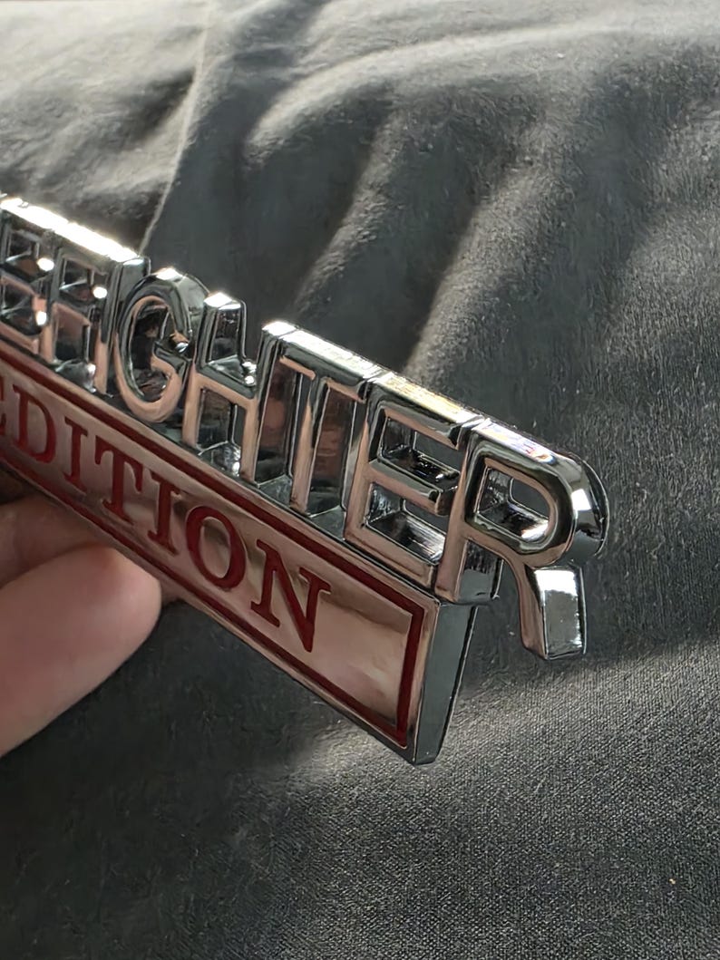 以下が含まれることがあります： 「FIGHTER」の文字が浮き彫りになったクロム仕上げのエンブレム。赤い文字で「EDITION」と書かれています。グレーの背景にエンブレムが配置されています。