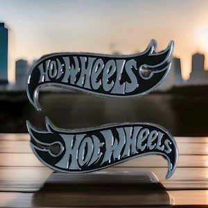 May include: Two black and silver Hot Wheels logo emblems. The word "Hot Wheels" is written in a stylized font within a black, curved emblem. The emblems are displayed on a reflective surface with a blurred cityscape background.