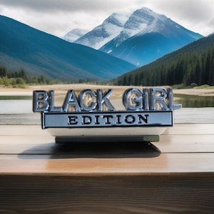 May include: A chrome emblem reading "BLACK GIRL EDITION" on a white and silver base. The emblem is set against a scenic backdrop of mountains, a lake, and trees, resting on a wooden surface.