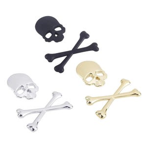 May include: A selection of skull and crossbones charms in black, silver, and gold. The charms have different finishes, including matte and metallic. The skulls have eye and nose sockets. The crossbones are crossed at the centre.