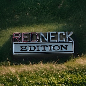Puede incluir: Un emblema cromado y rojo "REDNECK EDITION", sobre un fondo de hierba verde. El texto está en negrita, con la palabra "REDNECK" en letras rojas. El emblema tiene forma rectangular.