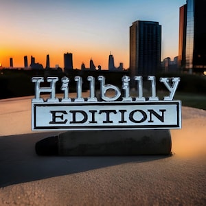 May include: A chrome "Hillbilly Edition" emblem with black text, set against a cityscape at sunset. The rectangular emblem has a black border and is mounted on a dark, cylindrical object.