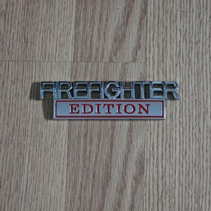 以下が含まれることがあります： 「FIREFIGHTER EDITION」の文字が入ったクロムメッキの金属製エンブレム。「FIREFIGHTER」の文字は、赤い背景の上に「EDITION」という文字の上に盛り上がったブロック体のフォントで表示されています。エンブレムは木製の表面にあります。