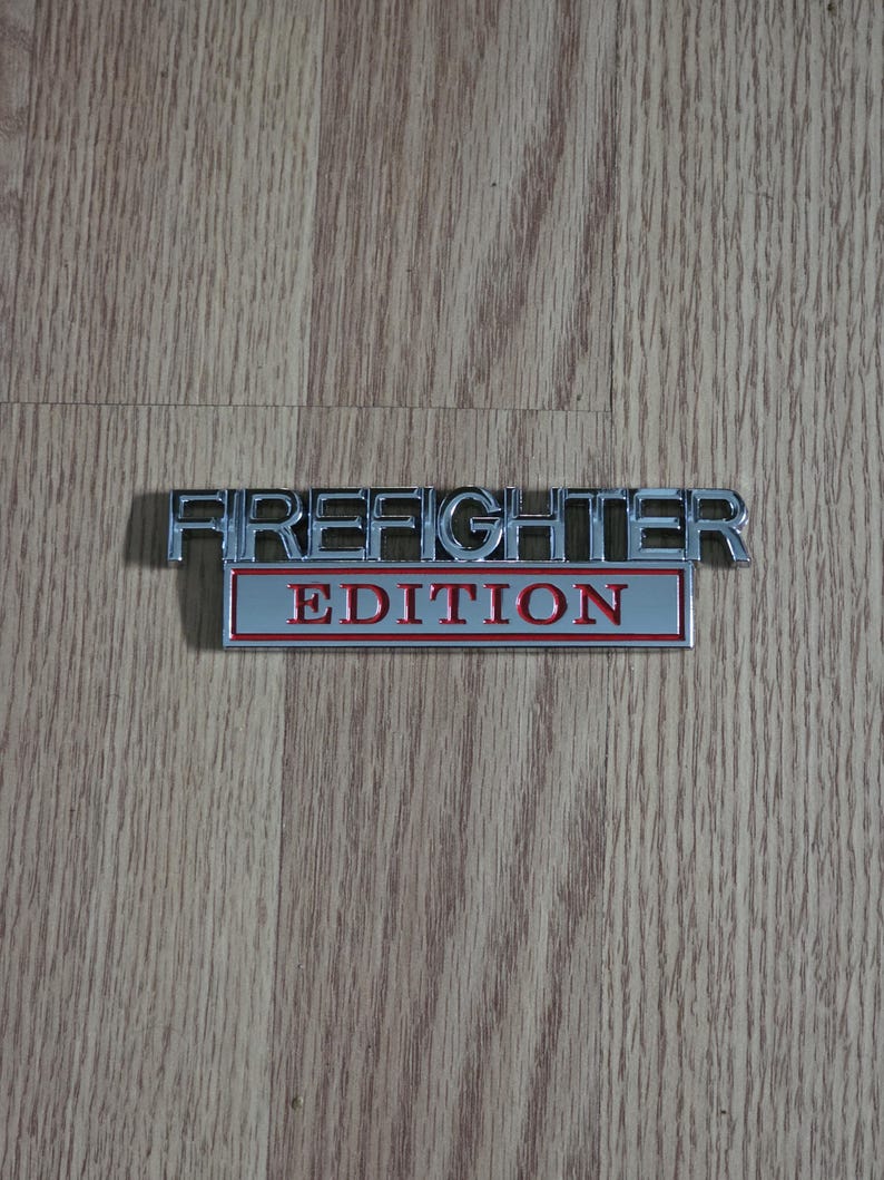 以下が含まれることがあります： 「FIREFIGHTER EDITION」の文字が入ったクロムメッキの金属製エンブレム。「FIREFIGHTER」の文字は、赤い背景の上に「EDITION」という文字の上に盛り上がったブロック体のフォントで表示されています。エンブレムは木製の表面にあります。