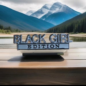 May include: A silver and black emblem reading "BLACK GIRL EDITION" is set against a backdrop of mountains and a lake. The emblem is positioned on a wooden surface.