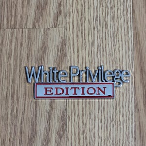 May include: A silver metal emblem with the words "White Privilege" in capital letters. Below, a rectangular plaque with the word "EDITION" in red letters on a gray background.