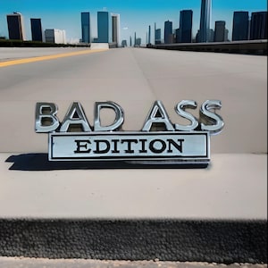 May include: Chrome "BAD ASS EDITION" emblem. The emblem is set against a light-coloured surface, with a cityscape in the background under a blue sky. The text is in a bold, sans-serif font.