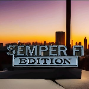 May include: A chrome emblem displaying "SEMPER FI EDITION" set against a sunset. The emblem is fixed on a dark surface, with a cityscape silhouette in the background.