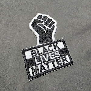 May include: A black and white embroidered patch featuring a raised fist above a rectangular sign. The sign reads "BLACK LIVES MATTER" in bold, white capital letters against a black background.