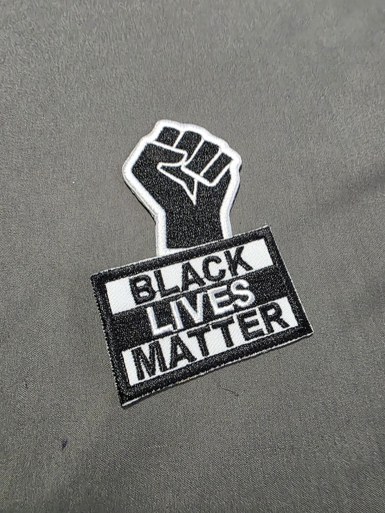 May include: A black and white embroidered patch featuring a raised fist above a rectangular sign. The sign reads "BLACK LIVES MATTER" in bold, white capital letters against a black background.