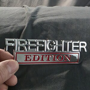 以下が含まれることがあります： 「FIREFIGHTER」の文字がクロム色の金属製エンブレムにあり、その下に赤い文字で「EDITION」と書かれています。長方形のエンブレムは、おそらく車のアクセサリーです。
