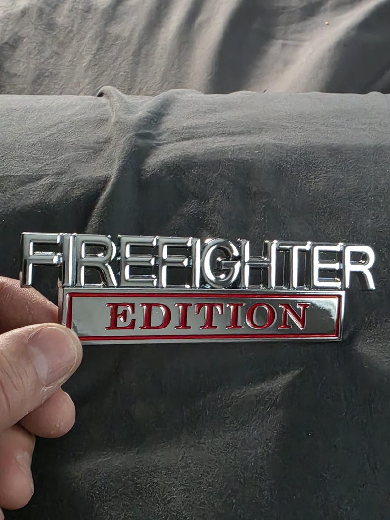以下が含まれることがあります： 「FIREFIGHTER」の文字がクロム色の金属製エンブレムにあり、その下に赤い文字で「EDITION」と書かれています。長方形のエンブレムは、おそらく車のアクセサリーです。