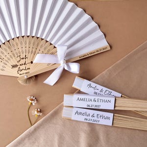 May include: A collection of personalized wedding fans. One large white paper fan with a wooden handle is open, and features the names "Jonh and Amelia" and the date "06.27.2027". Several smaller fans are also visible.
