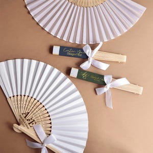May include: White folding fans with light wood ribs and white ribbon bows. One fan is open, showing the text "AE Amelia & Ethon 08.27.2027" on the wood. Two closed fans feature dark labels with "Custom Your Desgin/Text" and "AE Amelia & Ethon 08.27.2027".