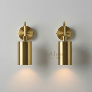 May include: Two matching brass wall sconces. Each sconce has a circular backplate, a cylindrical light fixture, and a curved arm. The sconces are mounted on a white wall and emit a warm, downward glow. The brand name "Alsa Craft Studio" is visible.