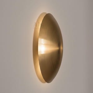Può includere: Applique da parete moderna in ottone con design a mezzaluna. L'applique ha una superficie liscia e riflettente che emette una luce calda. Montata su una parete di colore neutro, crea un'estetica minimalista ed elegante.