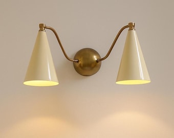 May include: A wall-mounted light fixture with two cream-colored, cone-shaped shades. The fixture has a brass-colored metal frame and a round base. The light is on, casting a warm glow.