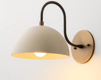 May include: Cream-colored dome-shaped wall sconce with a dark brown curved arm and a round brass-colored base. The sconce has a warm, inviting glow from the exposed lightbulb. A stylish and functional lighting fixture.
