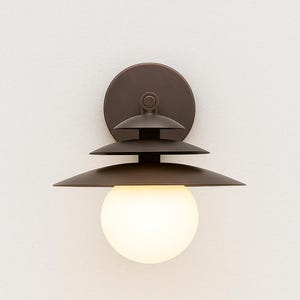 May include: A bronze wall sconce with a layered, geometric design. The fixture has a circular backplate, three tiered shades, and a spherical, frosted glass light. The sconce is designed for indoor lighting.