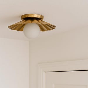 Puede incluir: Una lámpara de techo dorada con una pantalla festoneada y una bombilla redonda y blanca. La lámpara está montada en un techo blanco. La lámpara es un elemento decorativo.