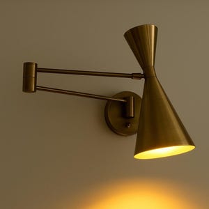 May include: A brass wall sconce featuring a conical shade and an adjustable arm. The light fitting is mounted on a circular base and emits a warm glow. The design is mid-century modern, with a retro aesthetic.