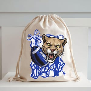 May include: A beige drawstring bag featuring a cougar head graphic with a blue football and the word "Cougars". The cougar is brown with yellow eyes and an open mouth. The football is blue and white.