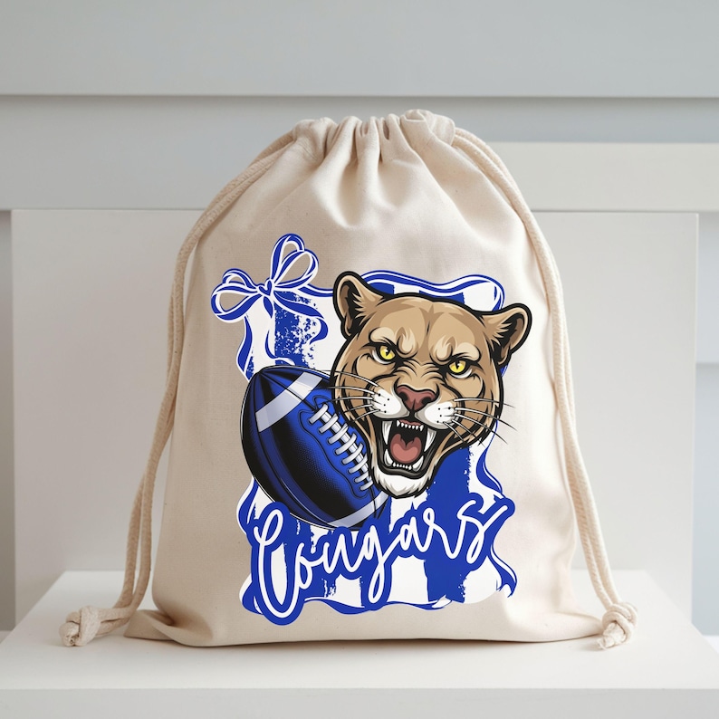 May include: A beige drawstring bag featuring a cougar head graphic with a blue football and the word "Cougars". The cougar is brown with yellow eyes and an open mouth. The football is blue and white.