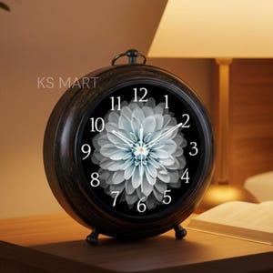 May include: A dark brown, round analog clock with a floral design on the face. The clock has a black face with white numbers and hands. The clock is on a wooden bedside table next to a lamp and an open book.