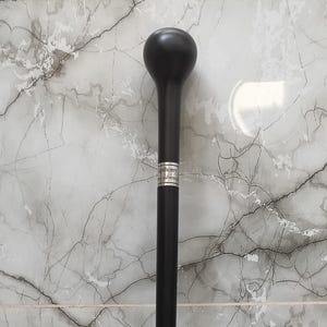 May include: A black walking cane with a rounded top and a slender shaft. The cane has a silver band near the top. The background is a light gray and white marble pattern.