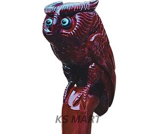 May include: A carved wooden owl figurine in a rich, dark red hue. The owl has detailed feather carvings, bright blue eyes, and is perched on a wooden base. The text "KS MART" is visible on the base.