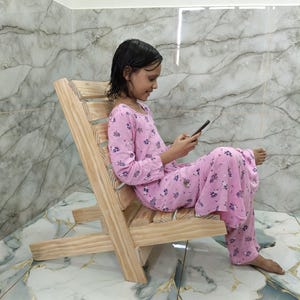 Puede incluir: Una silla de madera clara con asiento y respaldo de listones. La silla está inclinada hacia atrás, y una persona está sentada en ella, vistiendo un pijama rosa con un estampado floral, y usando un teléfono inteligente.
