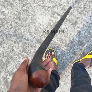 Peut inclure: Canne de marche noire avec une poignée incurvée et un pommeau en bois. La canne a une finition sombre et lisse. Le texte "KS MART" est visible sur la surface.