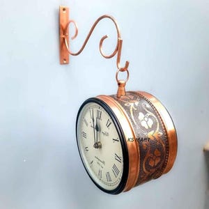 Puede incluir: Un reloj de pared de doble cara de color cobre con un diseño floral decorativo. La esfera del reloj tiene números romanos y manecillas negras. El reloj cuelga de un soporte decorativo de cobre.