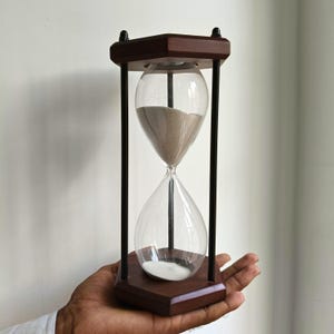 以下が含まれることがあります： 濃い茶色の木製フレームと黒い垂直サポートを備えた砂時計。 透明なガラス電球には、ベージュと白の砂が入っています。 砂時計は人の手で持たれています。