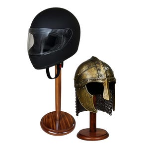 Könnte beinhalten: Zwei Helme auf Holzständern. Ein mattschwarzer Motorradhelm mit klarem Visier steht auf einem hohen Ständer. Daneben ein bronzefarbener mittelalterlicher Helm mit Kettenhemd, auf einem kürzeren Ständer. Beide Ständer haben einen runden Sockel.