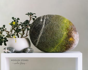 May include: A large, oval felted stone in shades of green, brown, and grey, with a white stripe. A white pot with a small plant and a few decorative stones are on a white shelf. The text "WOOLEN STONES" is visible.