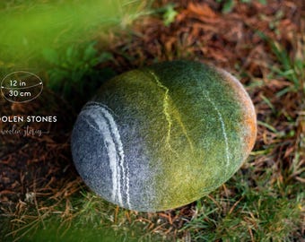 May include: A felted wool stone, approximately 30 cm in diameter, with a gradient of colours from blue-grey to green and yellow. The stone has a white stripe and is set against a backdrop of grass and foliage. The text "WOOLEN STONES" is visible.