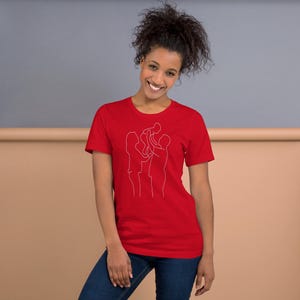 Puede incluir: Una camiseta roja con un dibujo lineal blanco de una familia con un niño. La camiseta es de manga corta y tiene cuello redondo. La modelo lleva vaqueros azules.