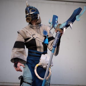 Puede incluir: Un disfraz que incluye un casco con máscara de gas azul y dorado, una chaqueta beige y negra con una borla azul y pantalones azul claro. El conjunto cuenta con un delantal azul oscuro, botas negras y un bastón azul grande con puntas plateadas.