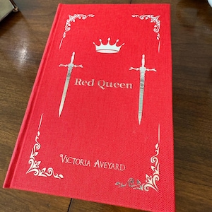 May include: A red hardcover book titled "Red Queen" by Victoria Aveyard. The cover features a silver crown, two swords, and decorative corner designs. The book is on a dark wooden surface.