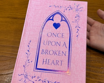 Puede incluir: Un libro rosa de tapa dura con el título "ONCE UPON A BROKEN HEART" en un marco decorativo. La portada presenta un corazón y detalles florales. El libro está sobre una superficie de madera.