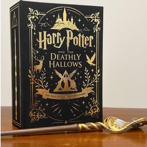 May include: A black book titled Harry Potter and the Deathly Hallows with gold foil illustrations of a castle, a stag, a bird, and the Deathly Hallows symbol. Beside the book lies a brown wand with a gold spiral handle and a clear crystal top.