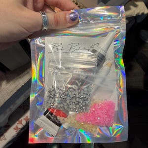 May include: A clear, iridescent zip bag containing various craft supplies. Inside are small bags of silver and pink rhinestones, a tube of adhesive, and other small items. The bag has the text "Bork Bedazzling SE!!".