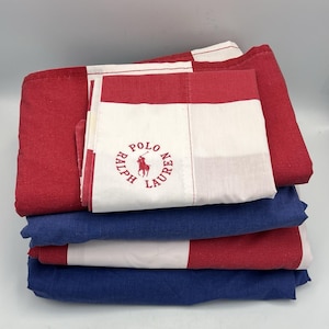 May include: A stack of folded bed linens in red, white, and blue. The top sheet features a red and white color block design with the "Polo Ralph Lauren" logo in red. The other sheets are solid blue and red and white color block.