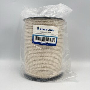 May include: A spool of River King macrame cord, wrapped in clear plastic. The label reads "Macrame Cord" and "220 Yards". The cord is a light beige color, and the spool is black.