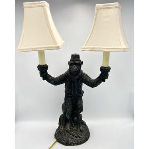 Puede incluir: Lámpara decorativa con una figura de mono sosteniendo dos pantallas de lámpara color crema. El mono lleva sombrero y chaleco, con acabado bronce. La lámpara tiene un diseño caprichoso y emite una luz cálida.