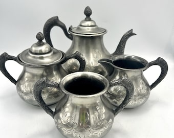May include: A four-piece silver-toned metal tea set comprising a teapot, a lidded sugar bowl, a creamer, and an open sugar bowl. Each piece features an etched floral pattern and dark, textured handles with a distinct, ornate design.