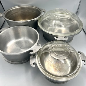 May include: A set of four silver-toned metal cooking pots with textured exterior finishes and smooth interior surfaces. Two pots feature clear glass lids with etched designs of a profile silhouette and crossed axes. Each pot has two side handles.