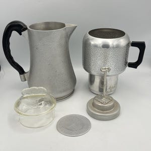 May include: A silver-toned aluminum coffee percolator set including a tall pitcher with a black handle, a cylindrical brewing chamber with a black handle, a clear glass lid with a scalloped edge, and a circular metal filter disc with a central stem.
